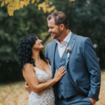 Elopement Wedding A couple stands close and smiles at each other under a tree with yellow leaves. The woman has black, curly hair and wears a sleeveless, white lace dress, while the man has short brown hair and wears a gray suit with a white shirt and a boutonniere. The background is filled with greenery. Elopements Inc